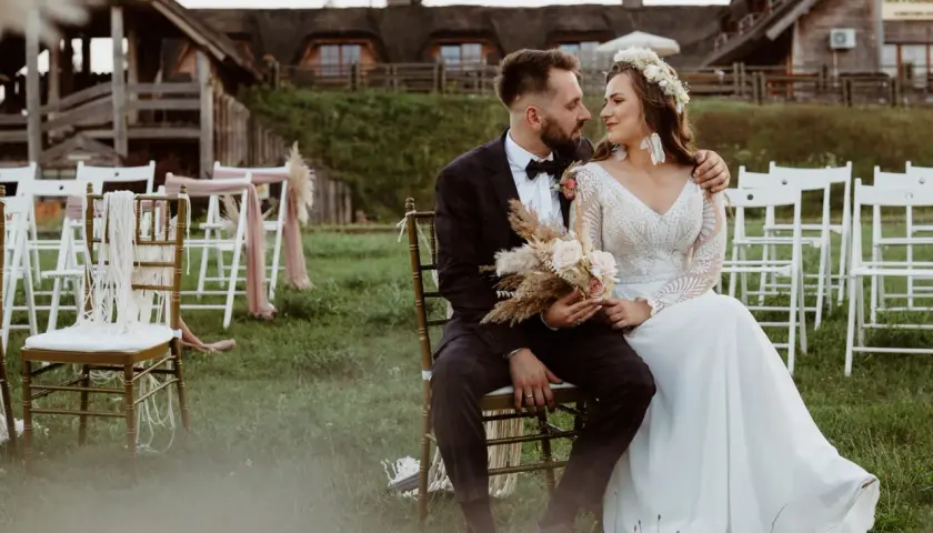 Para młoda siedzi na krzesłach na tle drewnianych budynków i białych krzeseł.